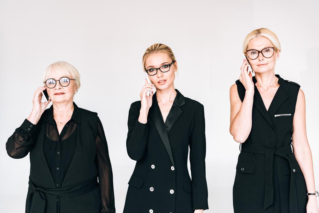 modische Geschäftsfrauen der dritten Generation in komplett schwarzen Outfits und Brille, die auf Smartphones sprechen, isoliert auf grau - Foto, Bild
