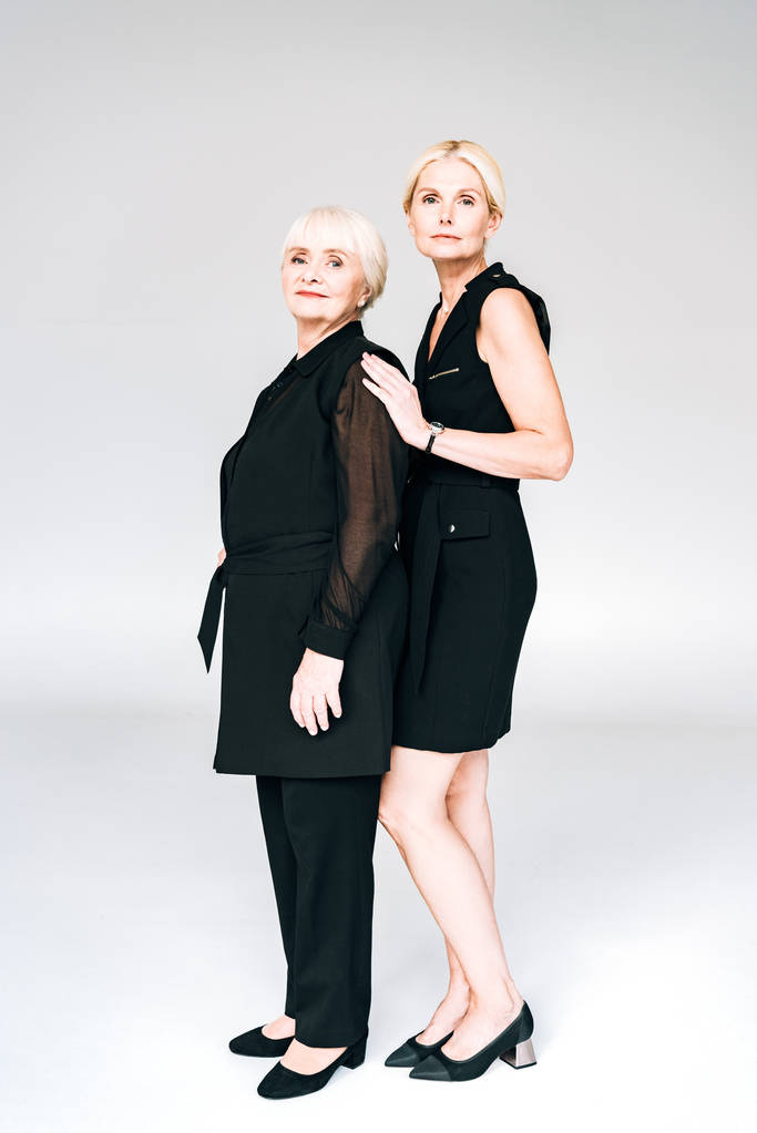 Ganzkörperansicht der eleganten blonden reifen Tochter und älteren Mutter in insgesamt schwarzen Outfits isoliert auf grau - Foto, Bild