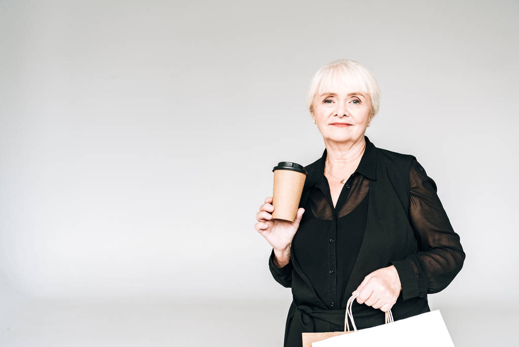 elegante Senior vrouw in zwarte outfit met boodschappentassen en koffie te gaan geïsoleerd op grijs - Foto, afbeelding