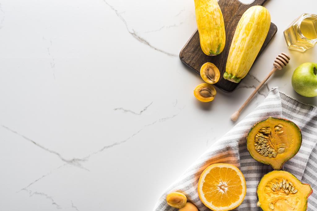 верхний вид полосатого полотенца с тыквой, абрикосами, цуккини и апельсином возле банки с медом и деревянной доской для резки на мраморной поверхности
 - Фото, изображение