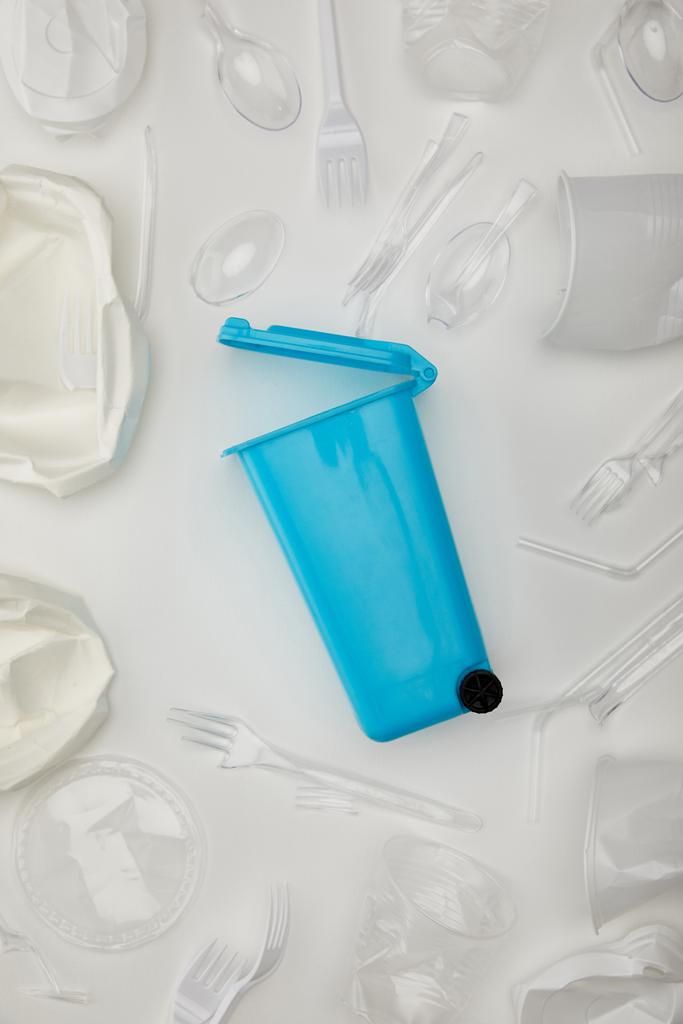 pohled na modrou odpadkový koš, zmačkané jednorázové šálky, vidličky, lžíce a talíře na bílém pozadí - Fotografie, Obrázek