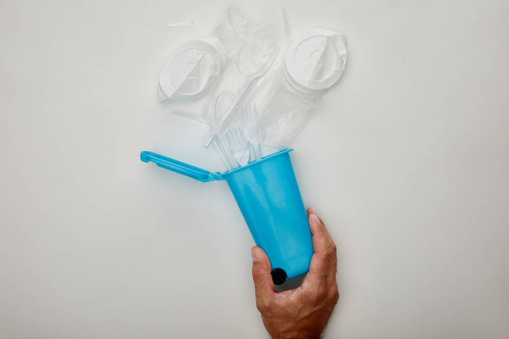 abgeschnittene Ansicht eines Mannes mit blauem Papierkorb mit zerknitterten Plastikbechern auf weißem Hintergrund - Foto, Bild