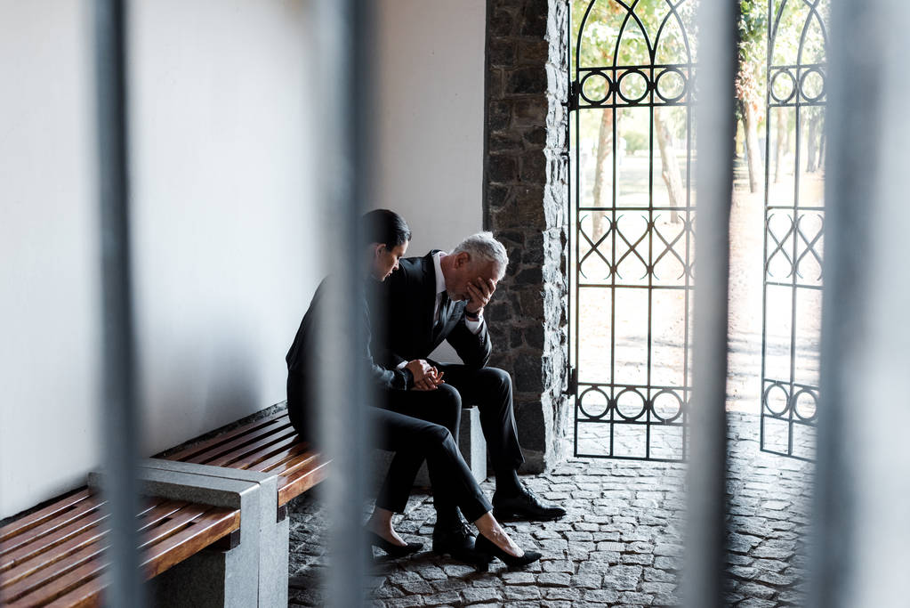 piękna kobieta siedzi na ławce w pobliżu smutnego starszego mężczyzny na cmentarzu  - Zdjęcie, obraz