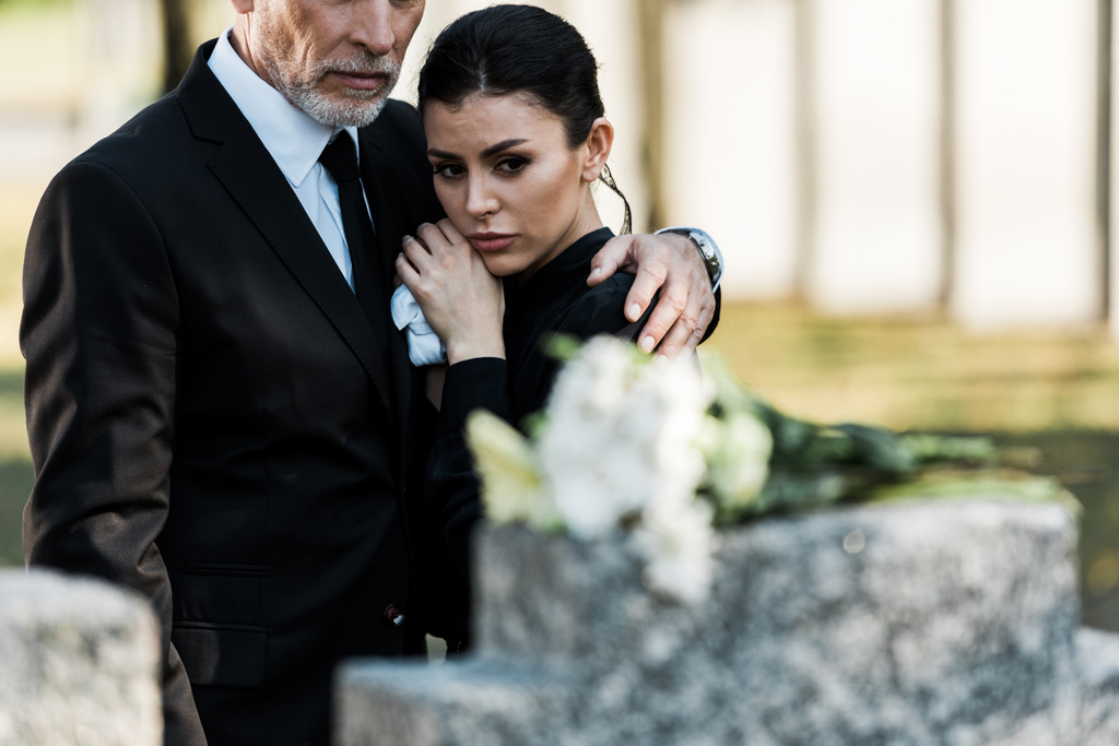 Selektiver Fokus eines bärtigen Mannes im Anzug, der attraktive Frau auf dem Friedhof umarmt  - Foto, Bild