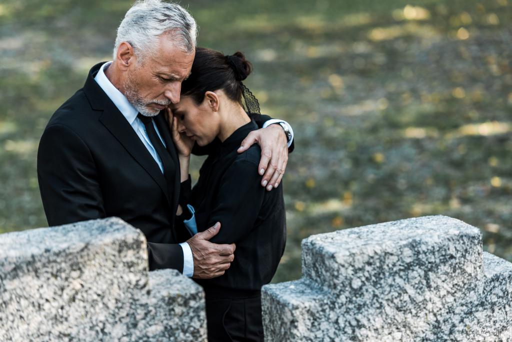 επιλεκτική εστίαση του γηραιότερος άντρας αγκαλιάζει γυναίκα κοντά σε τάφους  - Φωτογραφία, εικόνα