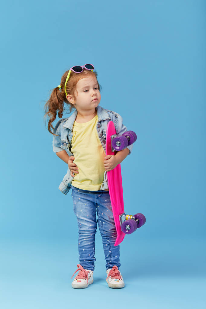Cool little toddler girl in denim clothes holds a skateboard and funny looking at the camera isolated on blue background - Photo, Image