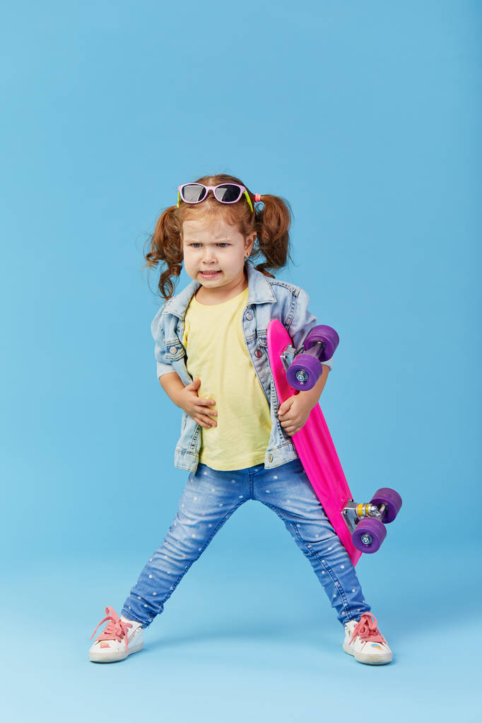 Stylowe fajne poważne małe dziecko dziewczyna z deskorolka na niebieskim tle. pokazując kciuk w dół niezadowolony. Miejsce na tekst - Zdjęcie, obraz