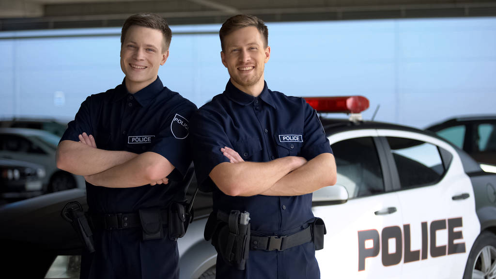 Lächelnde Polizisten mit überkreuzten Händen stehen vor Streifenwagen im Hintergrund - Foto, Bild