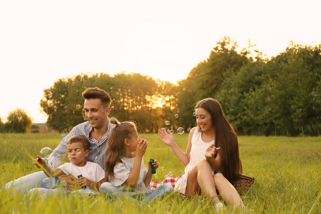 Šťastná rodina při západu slunce fouká v parku mýdlové bubliny. Letní piknik - Fotografie, Obrázek