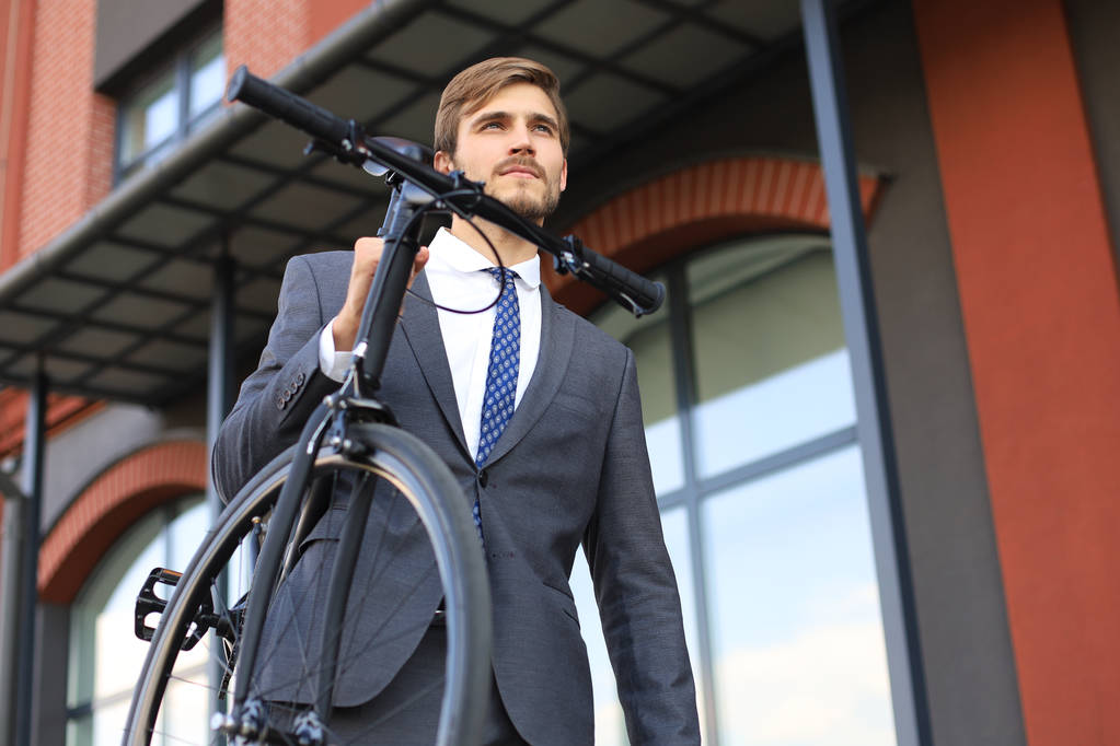 Guapo joven empresario llevando su bicicleta en el hombro mientras camina al aire libre
. - Foto, Imagen