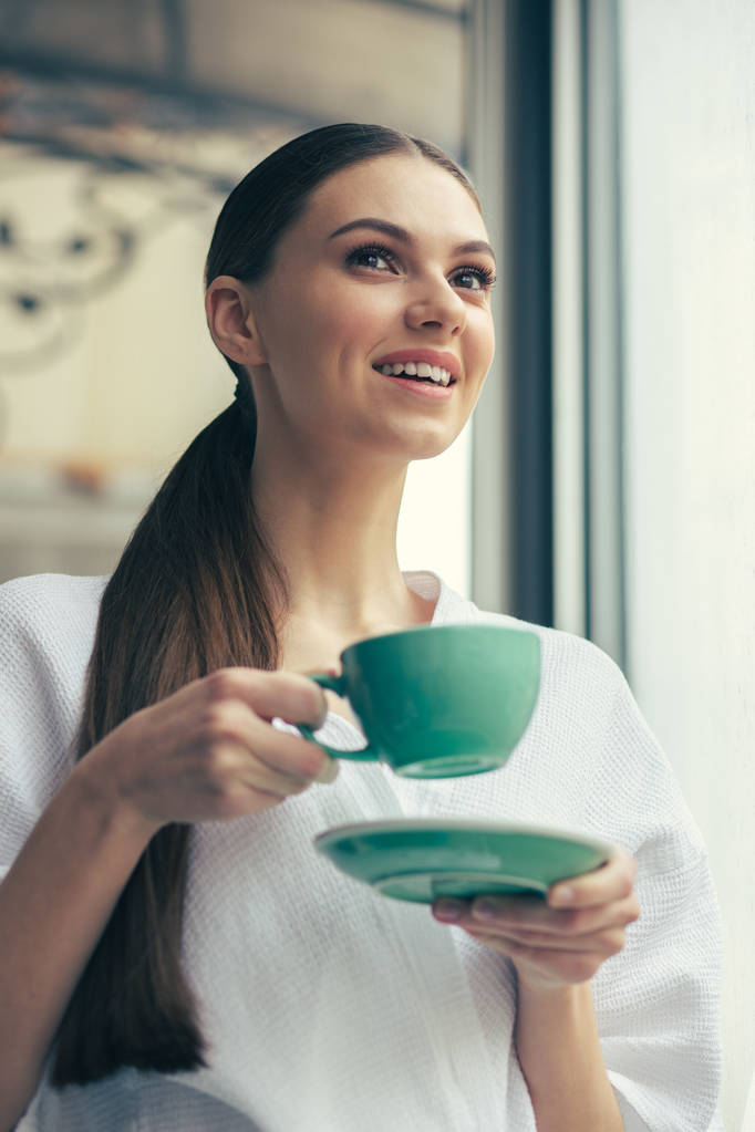 Περίεργη νεαρή γυναίκα με μακριά σκούρα μαλλιά στέκεται μόνη της με ένα φλιτζάνι τσάι και χαμογελά ενώ κοιτάζοντας την απόσταση - Φωτογραφία, εικόνα