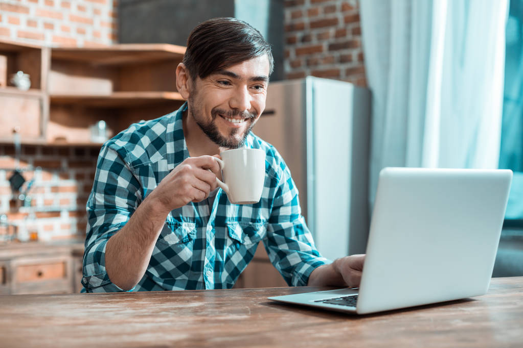 Lezzetli bir içecek. Mutlu pozitif akıllı adam dizüstü önünde oturan ve evden çalışırken çay sahip - Fotoğraf, Görsel