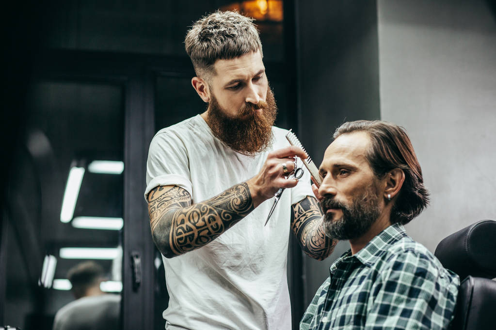 Középkorú férfi kockás ing ül a fodrászat, és keresi a nyugalmat, miközben tapasztalt Borbély, hosszú szakállú álló közelében és fésülje a haját - Fotó, kép
