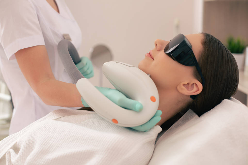 Cosmetologist experiente que está perto da mulher nova e conduzindo a remoção do cabelo do laser quando a mulher que usa óculos escuros
 - Foto, Imagem