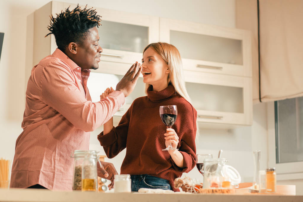陽気な若い女性は、彼女のボーイフレンドが彼女の鼻にいくつかの小麦粉を置いている間、ワインのグラスと笑いながら台所に立って - 写真・画像