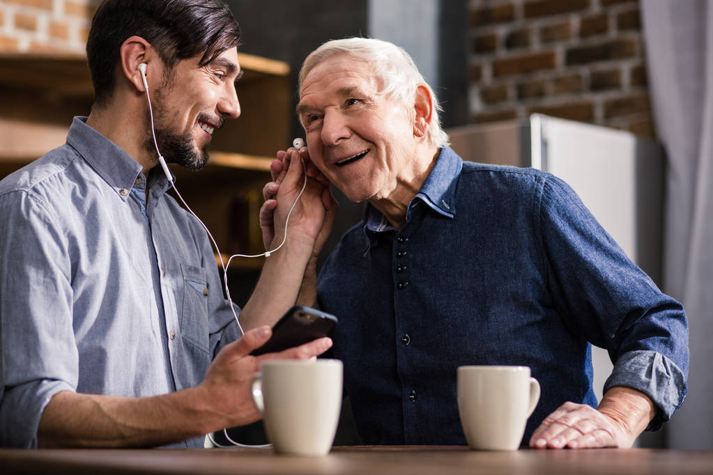 Χαίρομαι που σας μιλάω. Χαρούμενο ηλικιωμένο άνθρωπο χρησιμοποιώντας smartphone, ενώ αναπαύεται με το γιο του στην κουζίνα - Φωτογραφία, εικόνα
