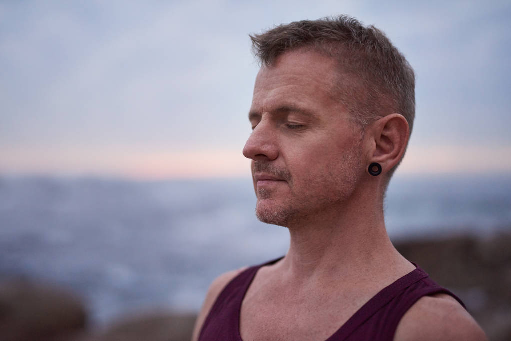 Ajuste o homem silenciosamente meditando enquanto está de pé com os olhos fechados pelo oceano durante a prática de ioga
 - Foto, Imagem