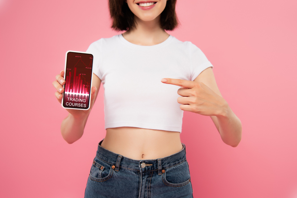 обрізаний вид дівчини, що вказує пальцем на смартфон із додатком торгових курсів ізольовано на рожевому
 - Фото, зображення
