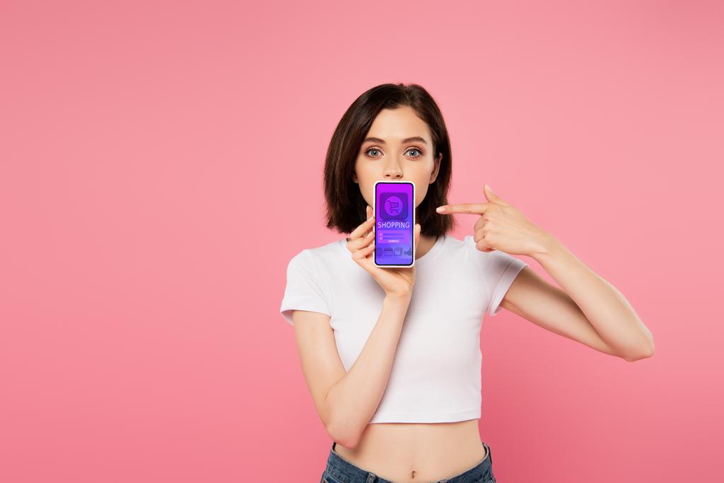 chocado chica apuntando con el dedo en el teléfono inteligente con la aplicación de compras aislado en rosa
 - Foto, imagen