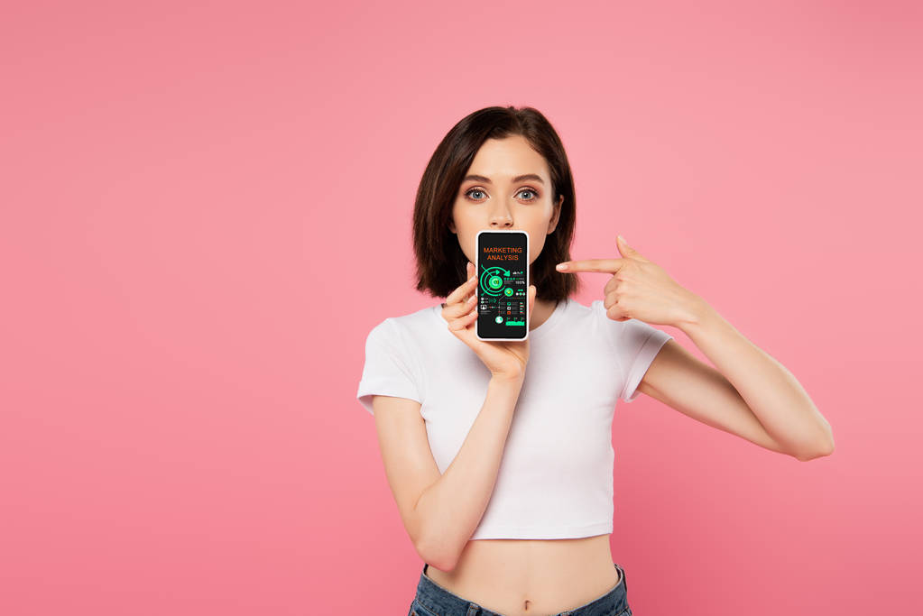 geschokt meisje wijzend met de vinger op de smartphone met marketing analyse geïsoleerd op roze - Foto, afbeelding