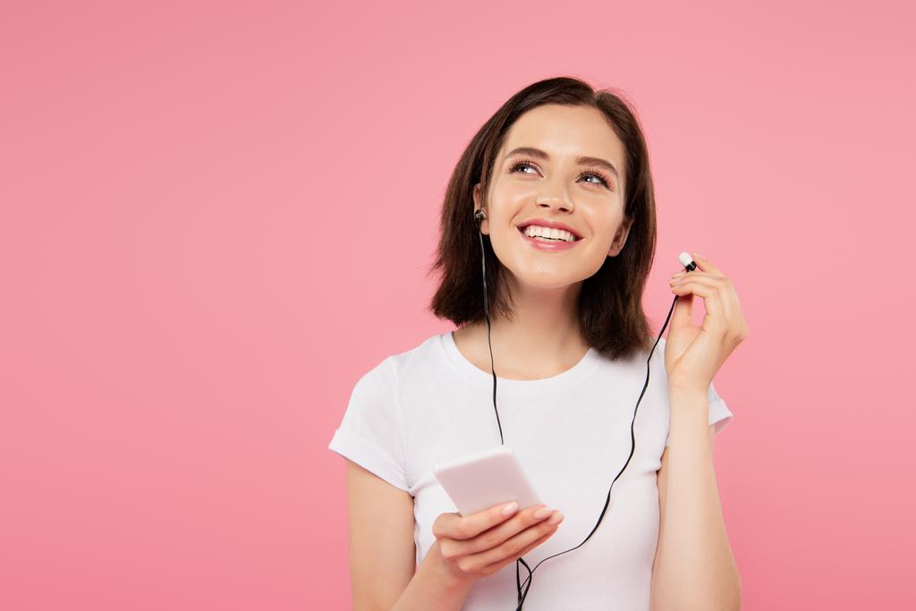 chica sonriente soñadora escuchando música en auriculares con teléfono inteligente aislado en rosa
  - Foto, imagen