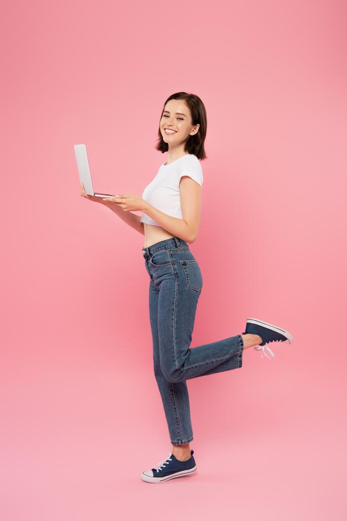 Вид в полный рост улыбающейся симпатичной девушки, позирующей с ноутбуком изолированной на розовом фоне
 - Фото, изображение