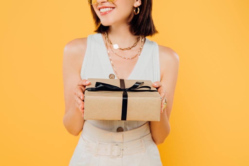 обрізаний вид усміхненої елегантної жінки, що тримає подарункову коробку ізольовано на жовтому
 - Фото, зображення