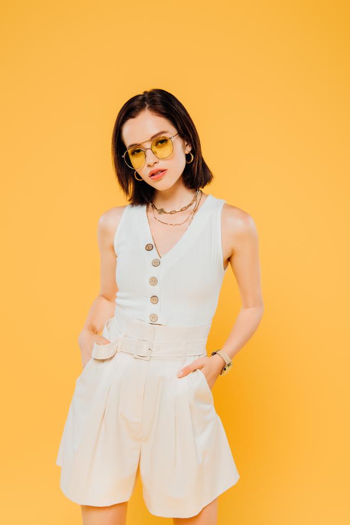 stylish woman in sunglasses posing with hands in pockets isolated on yellow - Photo, Image