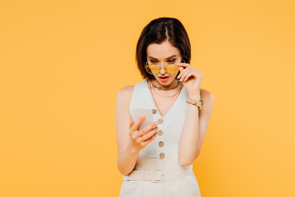 geschokt vrouw in zonnebrillen op zoek naar smartphone en aanraken zonnebril geïsoleerd op geel - Foto, afbeelding