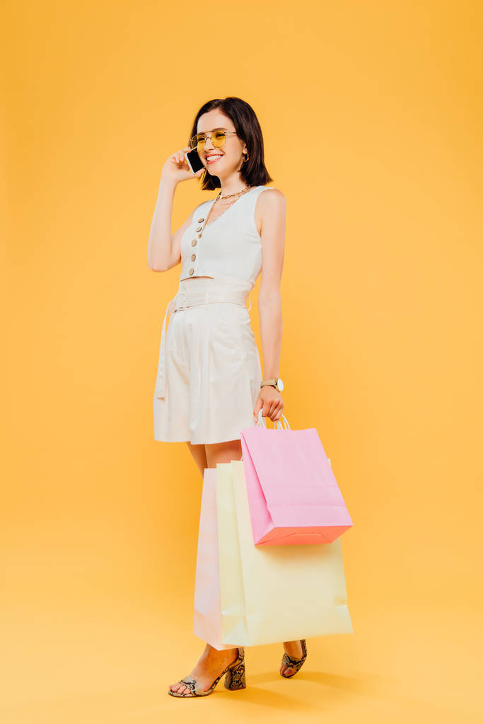 teljes hosszúságú kilátás mosolygó boldog nő napszemüveg gazdaságban bevásárló táskák és beszél okostelefon izolált sárga - Fotó, kép