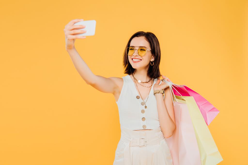 alışveriş çantaları ile güneş gözlüğü mutlu moda kız sarı izole akıllı telefonda selfie alarak - Fotoğraf, Görsel