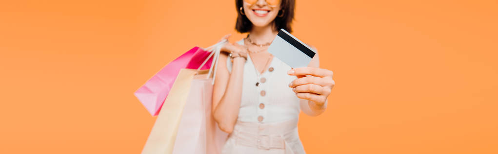 περικομμένη θέα του ευτυχισμένο κορίτσι με σακούλες για ψώνια που παρουσιάζουν πιστωτική κάρτα απομονώνονται σε πορτοκαλί - Φωτογραφία, εικόνα