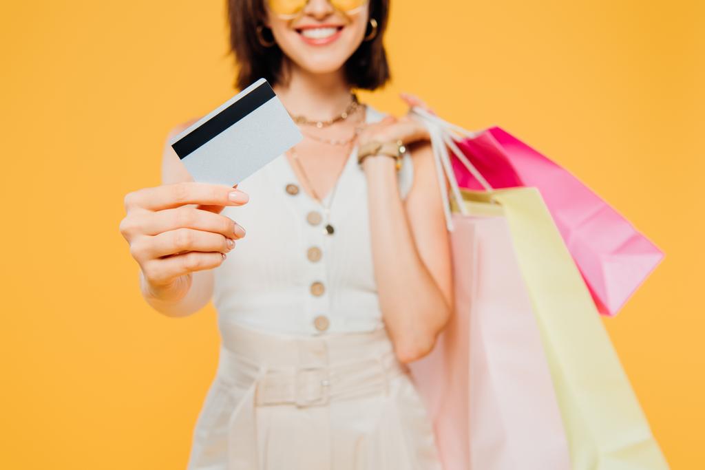 oříznutý pohled na šťastnou dívku s nákupními sáčky, které prezentuje kreditní kartu izolovanou na žluté - Fotografie, Obrázek