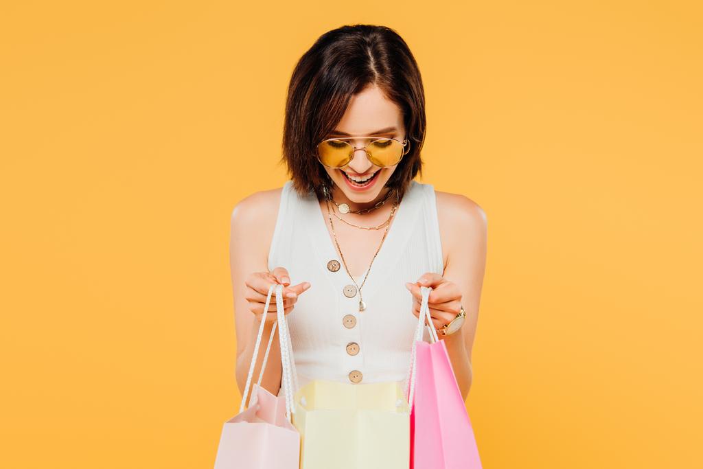 щаслива модна дівчина в сонцезахисних окулярах дивиться на сумки для покупок ізольовані на жовтому
 - Фото, зображення
