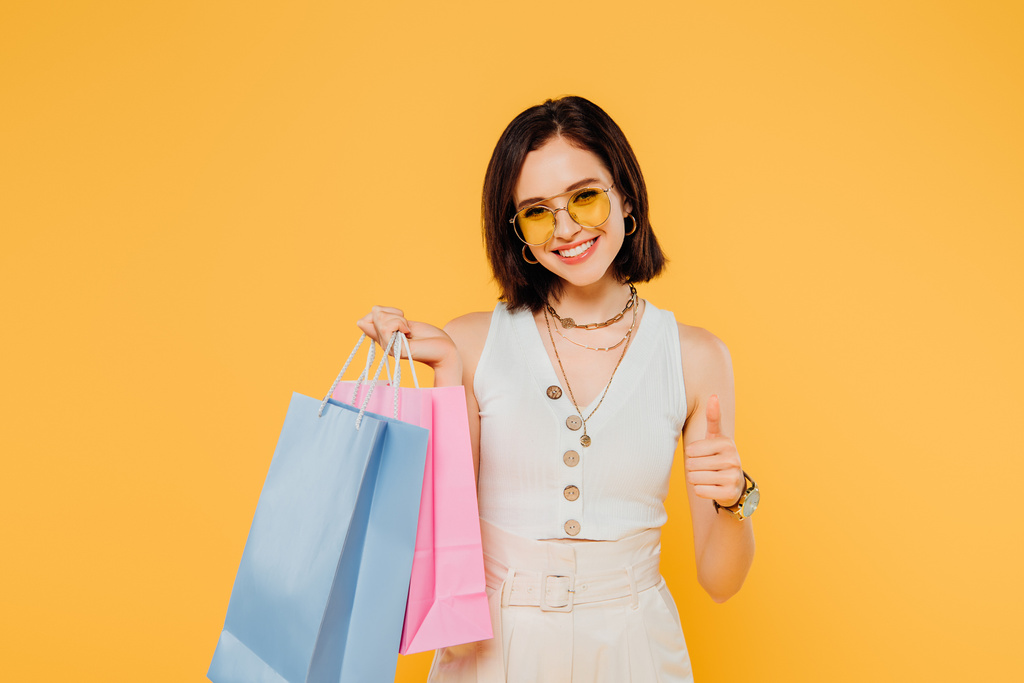 happy fashionable girl in sunglasses holding shopping bags and showing thumb up isolated on yellow - Photo, Image