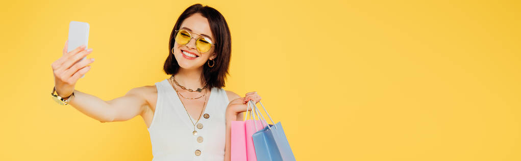 šťastná módní dívka v slunečních brýlích s nákupními sáčky, které si berou na sobě smartphone izolované na žluté - Fotografie, Obrázek