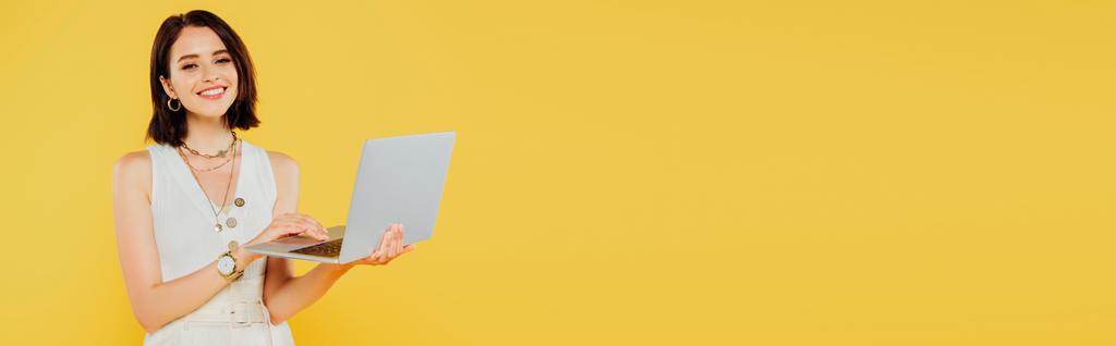 Panoramaaufnahme eines lächelnden eleganten Mädchens mit Laptop isoliert auf gelb - Foto, Bild