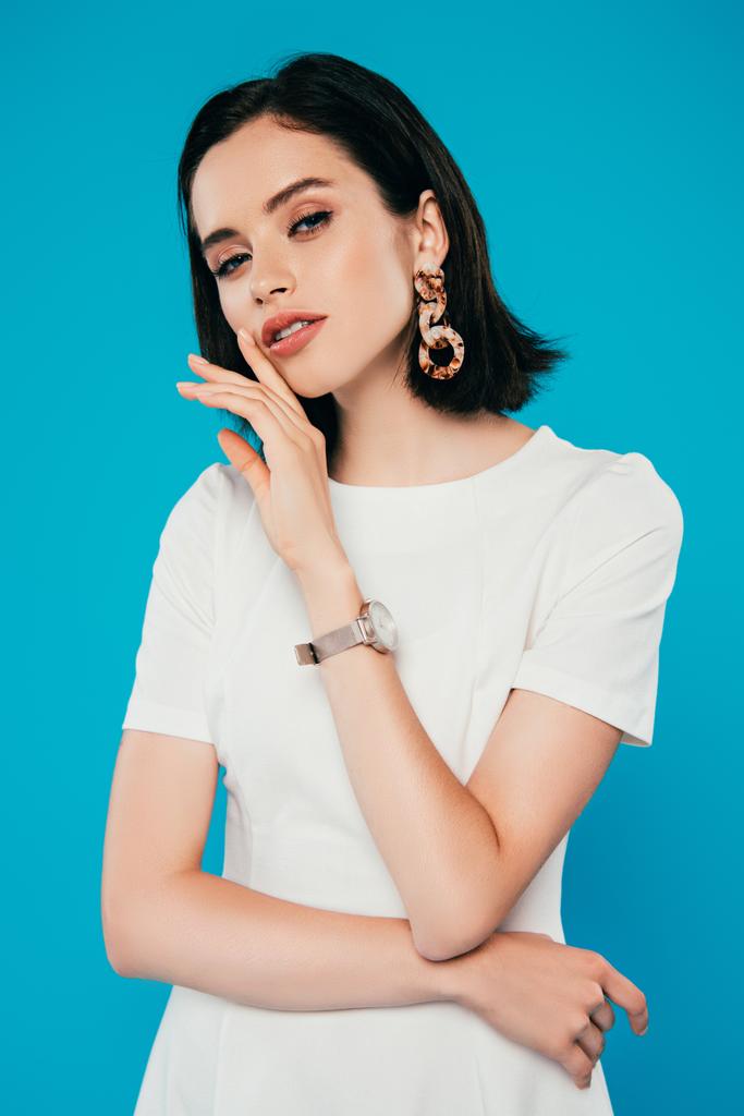 elegant woman in dress in earring and wristwatch with hand near face isolated on blue - Photo, Image