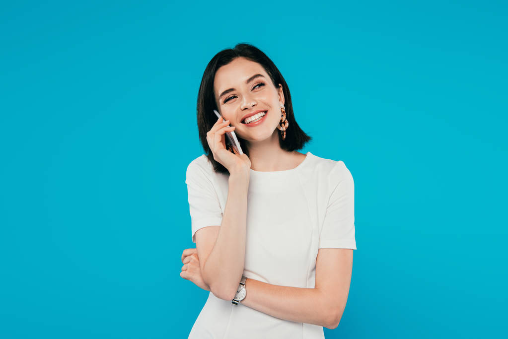 sonriente mujer elegante en vestido hablando en el teléfono inteligente y mirando hacia otro lado aislado en azul
 - Foto, imagen