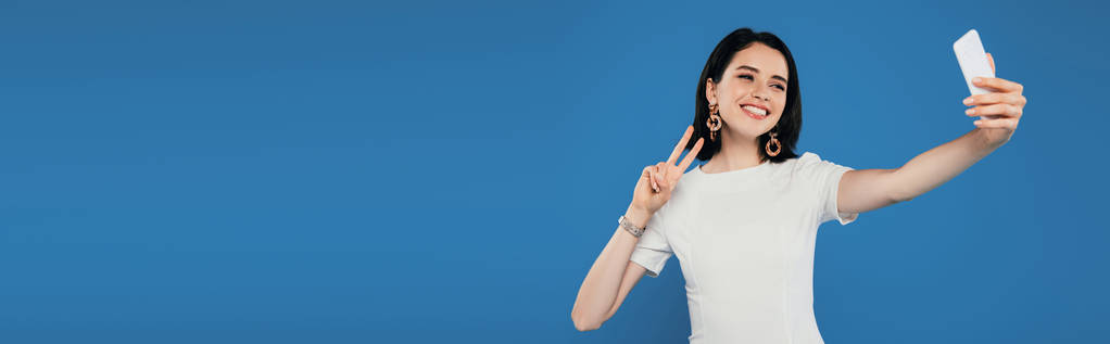 tiro panorâmico de sorrindo mulher elegante no vestido tomando selfie e mostrando sinal de paz isolado no azul
 - Foto, Imagem