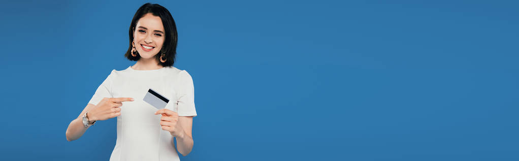 tiro panorâmico de mulher elegante sorridente no vestido apontando com o dedo para o cartão de crédito isolado no azul
 - Foto, Imagem