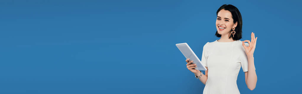 panoramiczny strzał uśmiechnięta elegancka kobieta trzyma cyfrowego tabletu i pokazując znak w porządku na niebiesko - Zdjęcie, obraz