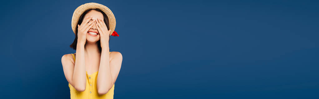 Uśmiechnięta dziewczyna w słomkowym kapeluszu z rękami na oczach wyizolowanych na niebiesko - Zdjęcie, obraz