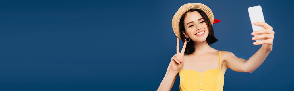 усміхнена дівчина в солом'яному капелюсі бере селфі на смартфон і показує знак миру ізольовано на синьому
 - Фото, зображення