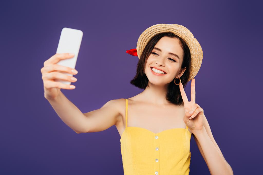 χαμογελαστή κορίτσι σε ψάθινο καπέλο παίρνοντας selfie στο smartphone και δείχνοντας το σημάδι της ειρήνης απομονωθεί σε μωβ - Φωτογραφία, εικόνα