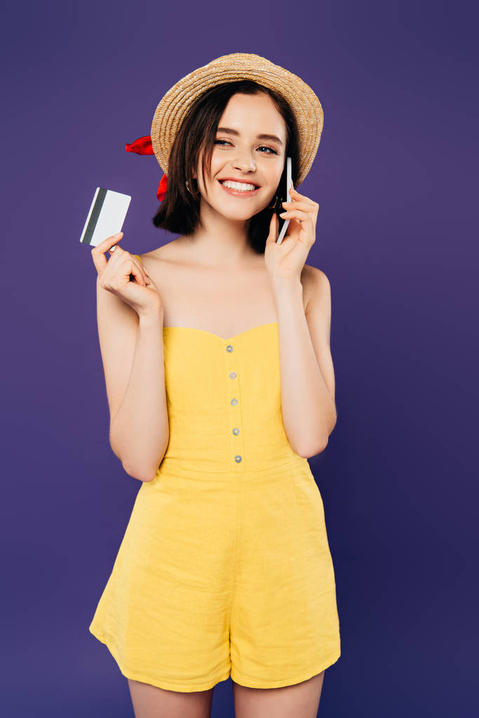 χαμογελαστή κορίτσι με ψάθινο καπέλο μιλάει στο smartphone και κατοχή πιστωτική κάρτα απομονωθεί σε μωβ - Φωτογραφία, εικόνα
