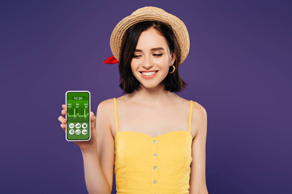 усміхнена дівчина в солом'яному капелюсі тримає смартфон із додатком охорони здоров'я ізольовано на фіолетовому
 - Фото, зображення