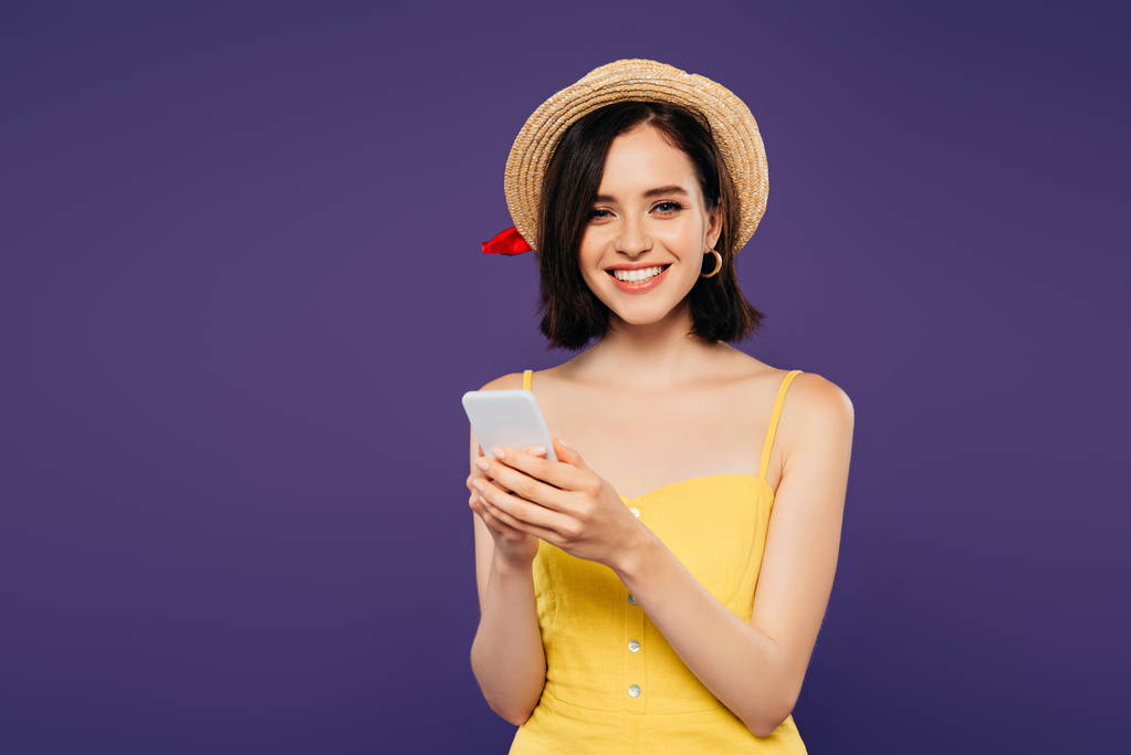 χαμογελαστή όμορφη κοπέλα σε ψάθινο καπέλο χρησιμοποιώντας smartphone απομονωθεί σε μωβ  - Φωτογραφία, εικόνα