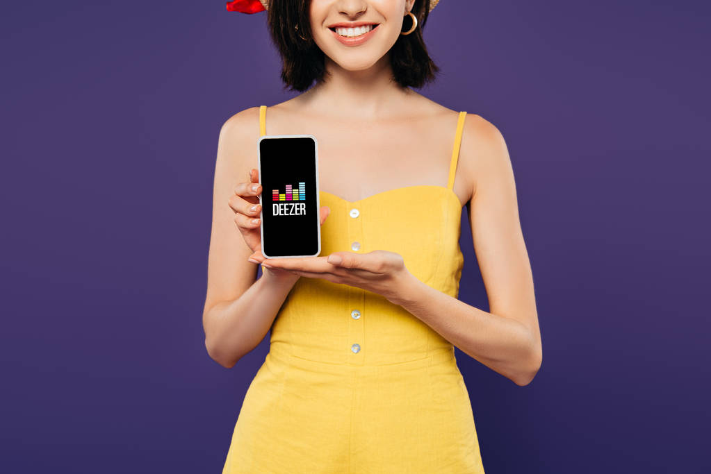 КИЕВ, Украина - 3 июля 2019 года: обрезанный вид улыбающейся девушки, держащей смартфон с приложением Deezer изолированным на фиолетовый
  - Фото, изображение
