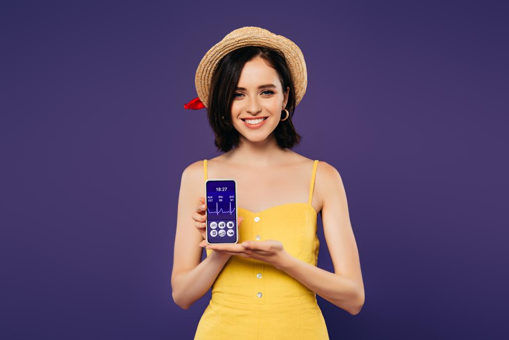 usmívající se krásná dívka v slaměném klobouku s smartphone se zdravotnickým aplikací izolovanými na purpurovém  - Fotografie, Obrázek